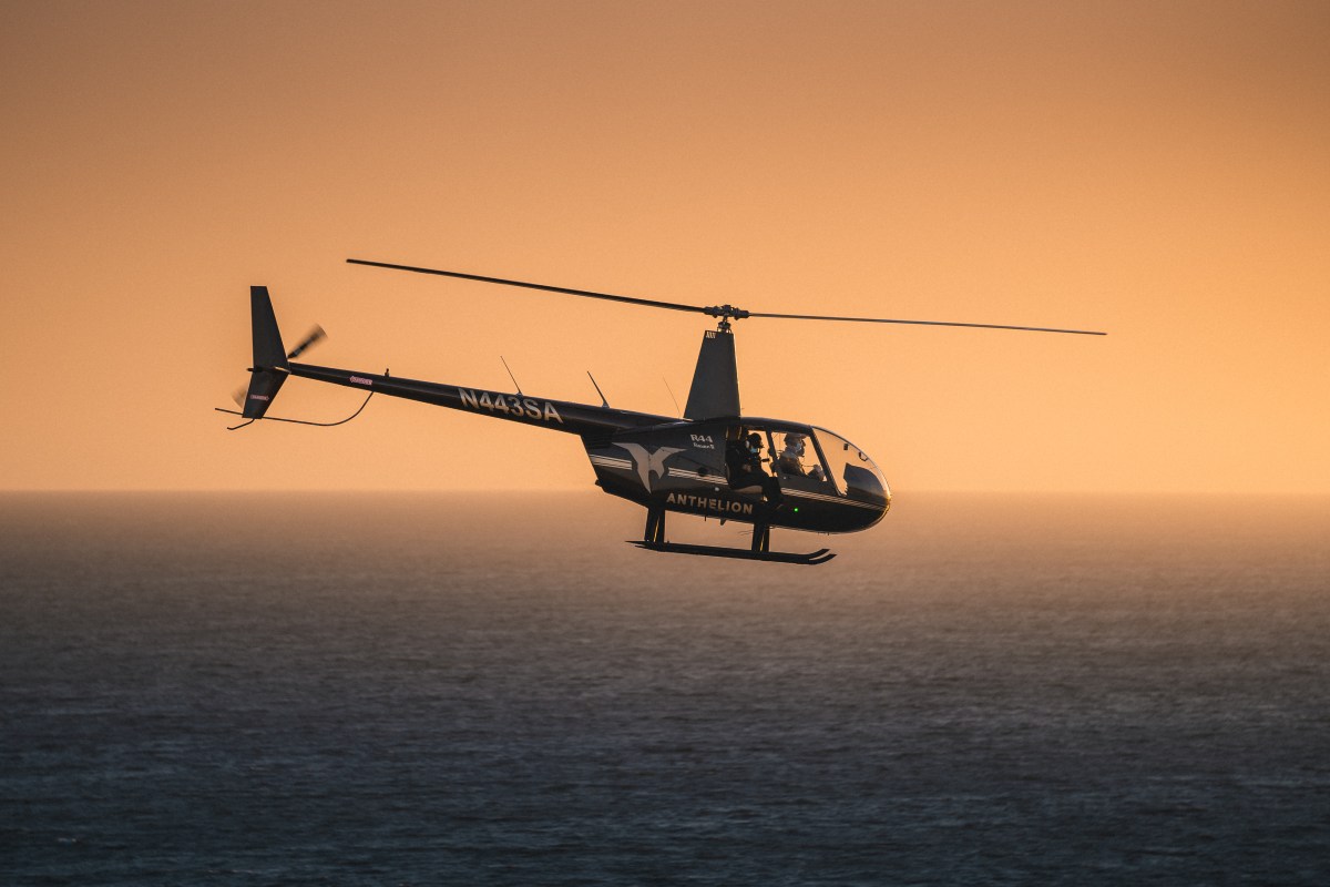 Helicopter flying over ocean at sunset with orange sky in background.