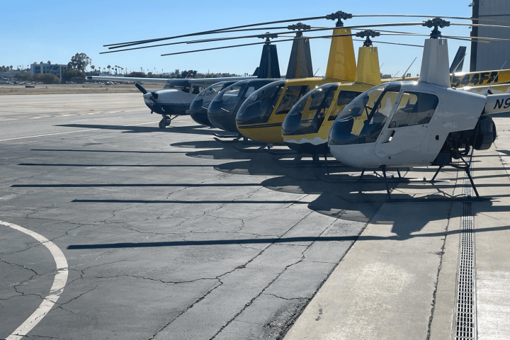 Seven helicopters lined up on an airport tarmac under clear skies.