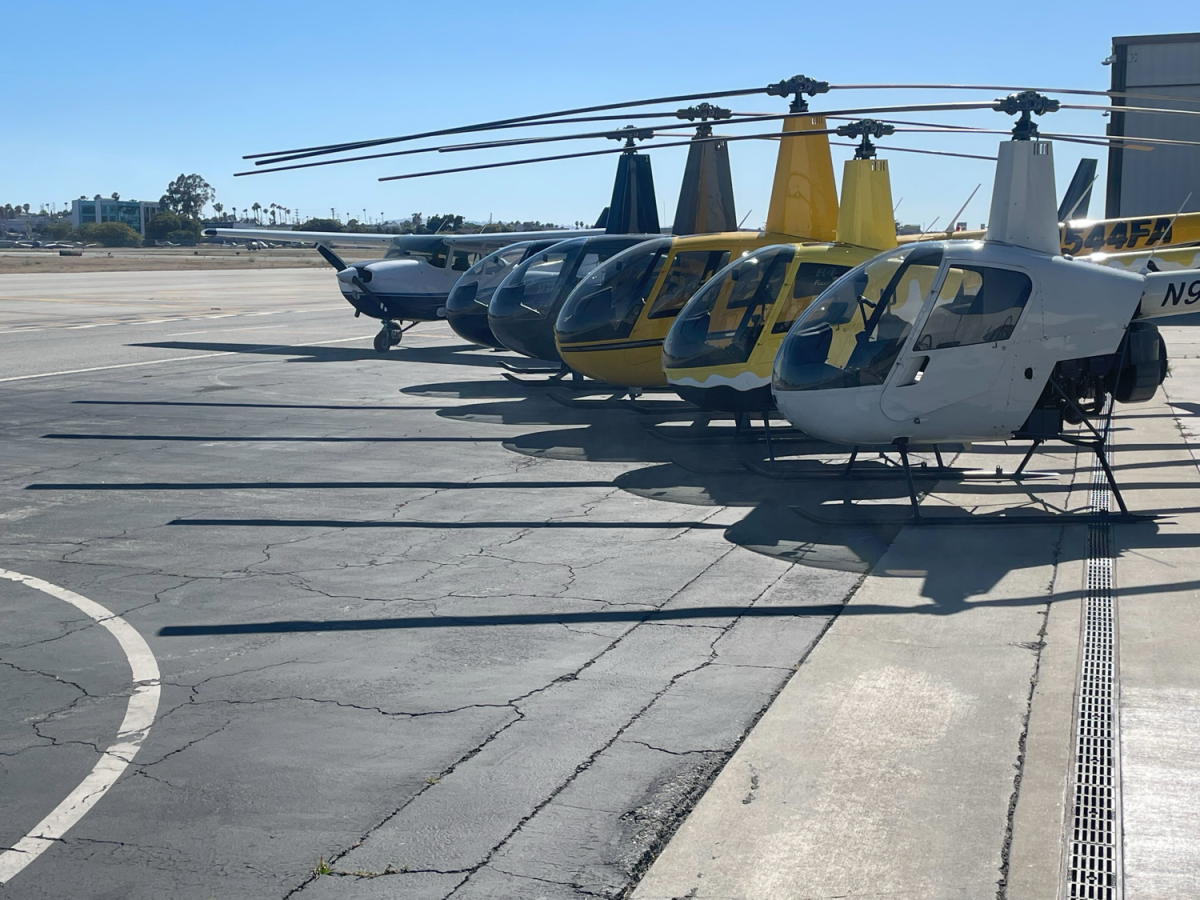 Seven helicopters lined up on an airport tarmac under clear skies.