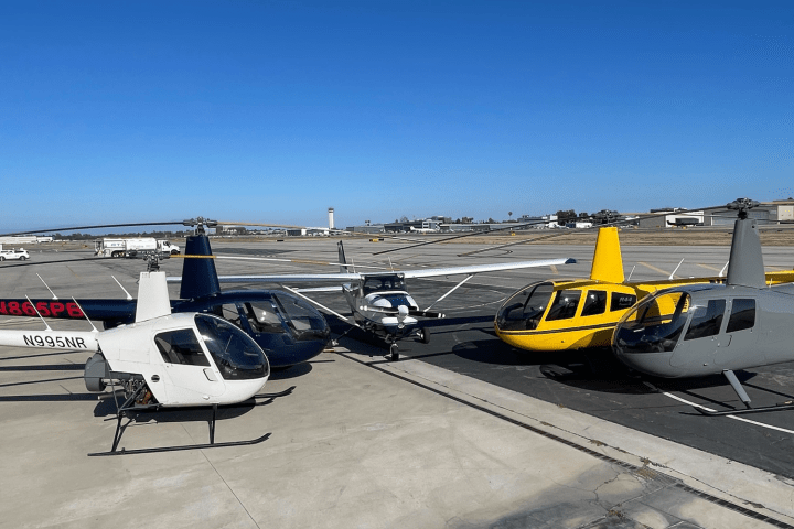 Four helicopters and a small plane parked on an airport tarmac under a clear blue sky.