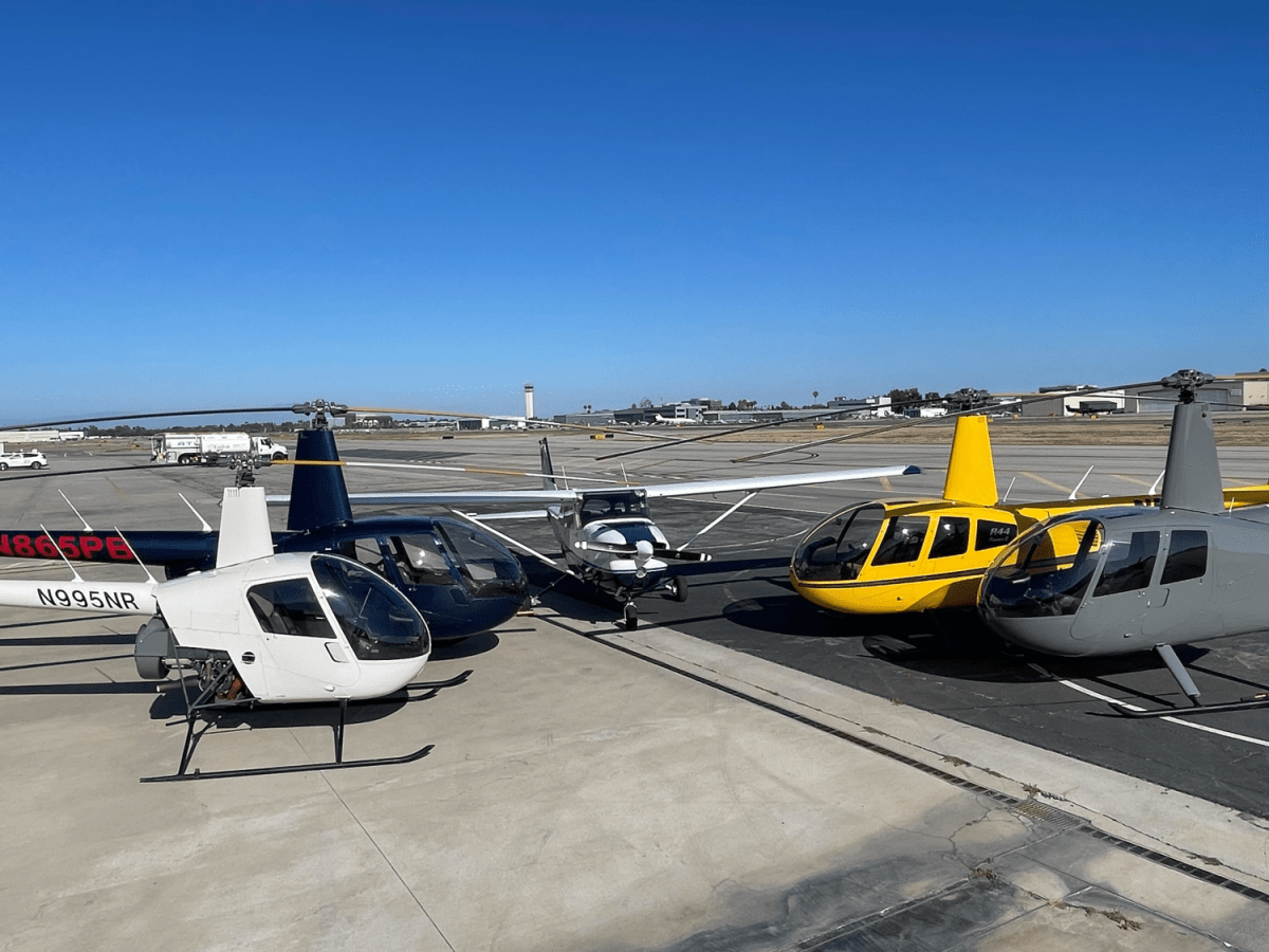 Four helicopters and a small plane parked on an airport tarmac under a clear blue sky.